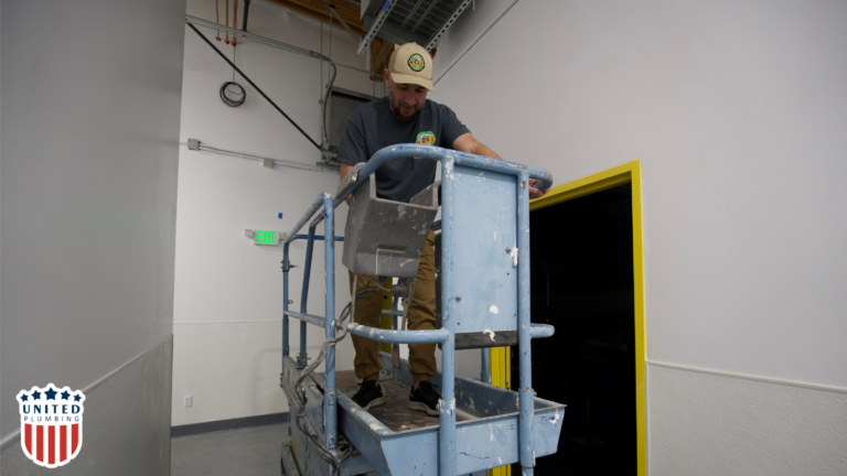 United Plumbing technician standing on a scissor lift inspecting pipes and plumbing system in a commercial building.