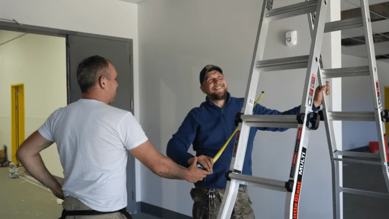Two tradesmen measuring near a ladder while discussing a project inside a building representing teamwork and communication in the trades industry.
